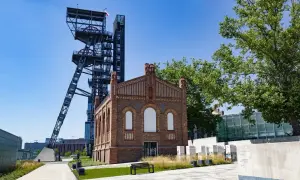 Dawna wieża szybu kopalnianego i ceglany budynek tworzą nietypowe atrakcje Katowice. Teren postindustrialny z zielenią i ławką.