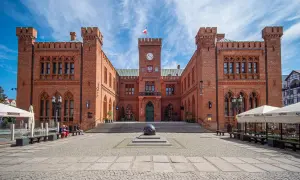 Rynek Kołobrzeg: imponujący ratusz z cegły, plac z kamienną rzeźbą i ogródki kawiarniane pod błękitnym niebem.