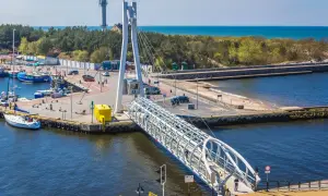 Port w Ustce z charakterystycznym mostem pieszym. W tle widać morze i las, a po lewej stronie cumują łodzie. Idealne miejsce na spacer przed wizytą w aquapark Ustka.