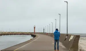 Mężczyzna w niebieskiej kurtce stoi na łebskiej promenadzie, patrząc na morze. W oddali widać latarnie morskie.