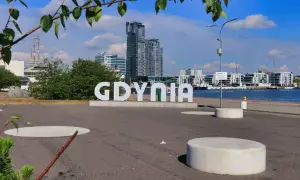Napis "GDYNIA" na tle nowoczesnych budynków i portu. Widoczny żaglowiec i błękitne niebo.