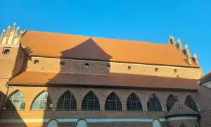 Zamek kapituły warmińskiej w Olsztynie, ceglana budowla z pomarańczowym dachem i łukowymi oknami, lśni w słońcu.