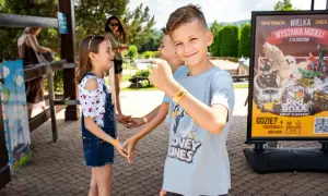 Uśmiechnięty chłopiec w parku rozrywki, obok niego dzieci. W tle plakat "Wielka wystawa modeli z klocków", który może zawierać informacje o **inwałd park cennik**.