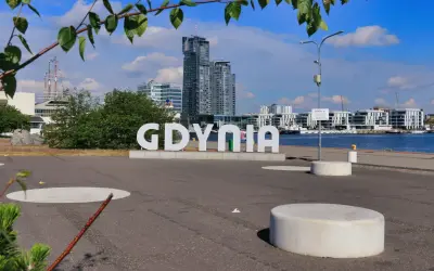 Napis "GDYNIA" na tle nowoczesnych budynków i portu. Widoczny żaglowiec i błękitne niebo.