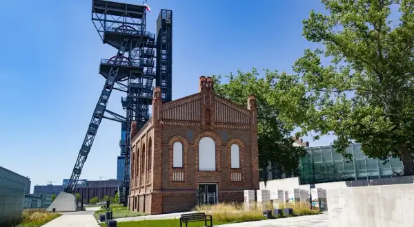 Dawna wieża szybu kopalnianego i ceglany budynek tworzą nietypowe atrakcje Katowice. Teren postindustrialny z zielenią i ławką.