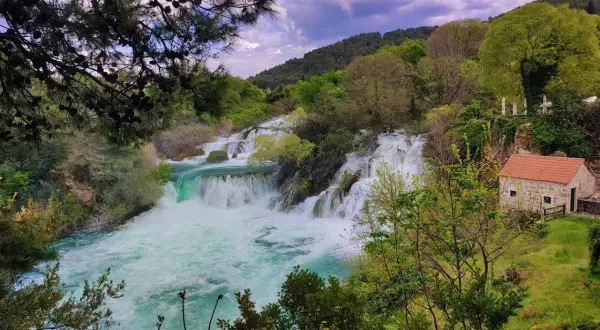 Zadar wodospad w Chorwacji, turkusowa woda spływa kaskadami wśród zieleni. Obok kamienny domek z czerwoną dachem.
