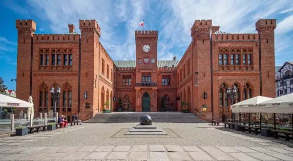 Rynek Kołobrzeg: imponujący ratusz z cegły, plac z kamienną rzeźbą i ogródki kawiarniane pod błękitnym niebem.