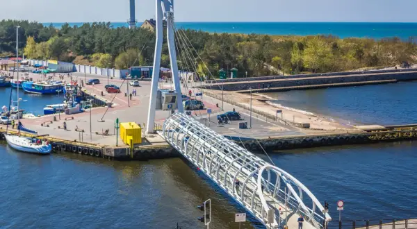 Port w Ustce z charakterystycznym mostem pieszym. W tle widać morze i las, a po lewej stronie cumują łodzie. Idealne miejsce na spacer przed wizytą w aquapark Ustka.