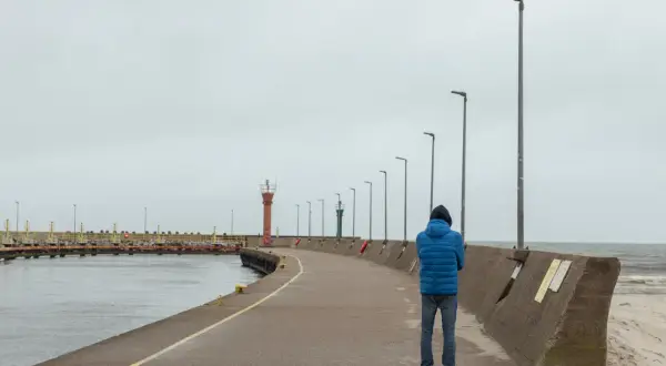Mężczyzna w niebieskiej kurtce stoi na łebskiej promenadzie, patrząc na morze. W oddali widać latarnie morskie.