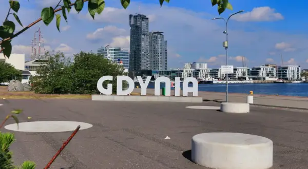 Napis "GDYNIA" na tle nowoczesnych budynków i portu. Widoczny żaglowiec i błękitne niebo.