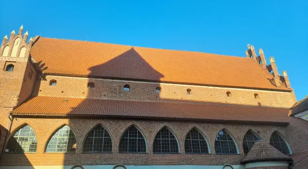 Zamek kapituły warmińskiej w Olsztynie, ceglana budowla z pomarańczowym dachem i łukowymi oknami, lśni w słońcu.