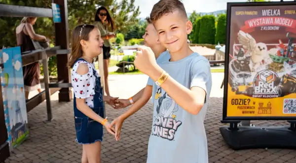 Uśmiechnięty chłopiec w parku rozrywki, obok niego dzieci. W tle plakat "Wielka wystawa modeli z klocków", który może zawierać informacje o **inwałd park cennik**.