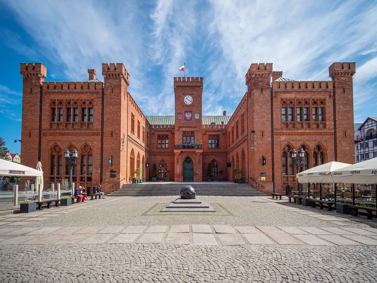 Rynek Kołobrzeg: imponujący ratusz z cegły, plac z kamienną rzeźbą i ogródki kawiarniane pod błękitnym niebem.