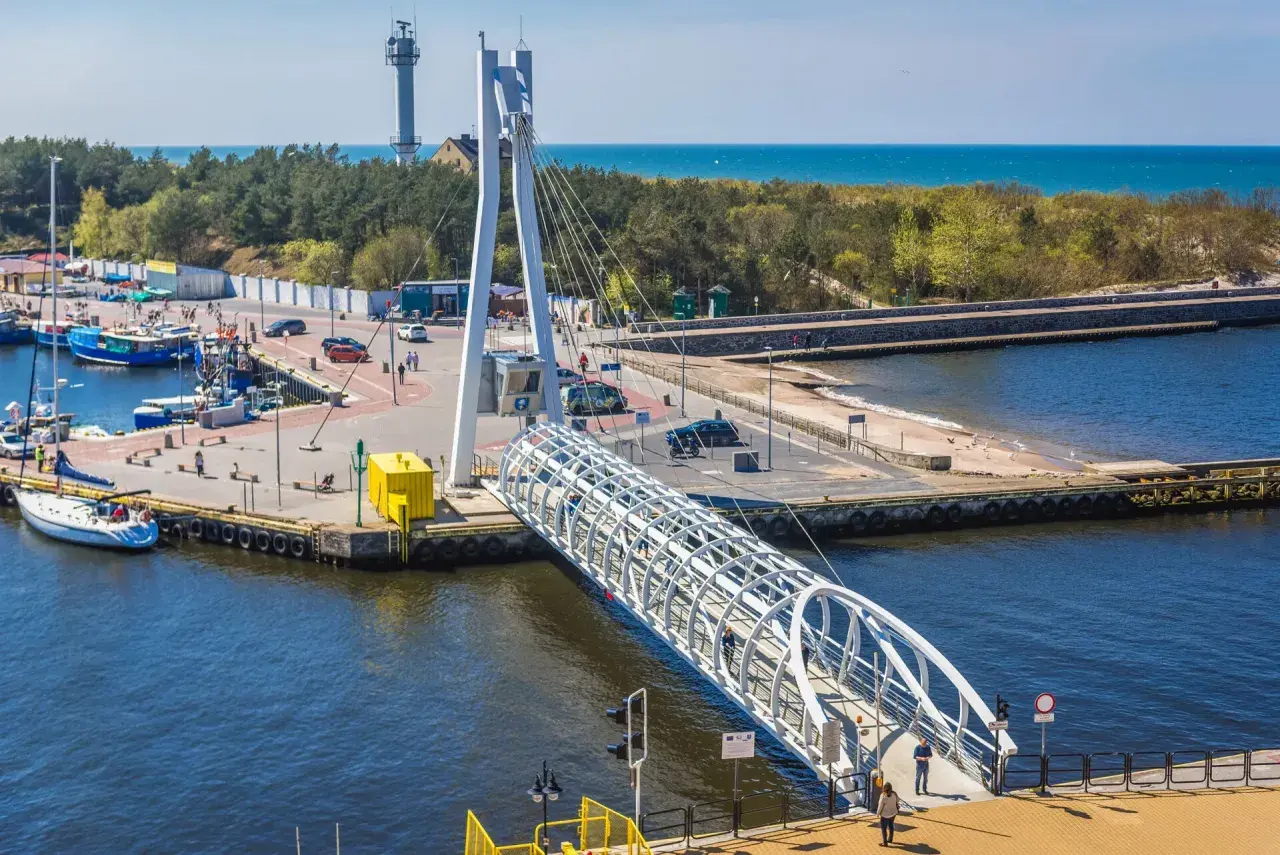 Port w Ustce z charakterystycznym mostem pieszym. W tle widać morze i las, a po lewej stronie cumują łodzie. Idealne miejsce na spacer przed wizytą w aquapark Ustka.