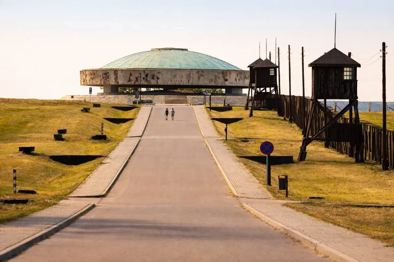 Droga prowadzi do Mauzoleum na terenie byłego obozu Majdanek. Dwie postacie idą w stronę monumentalnej budowli.