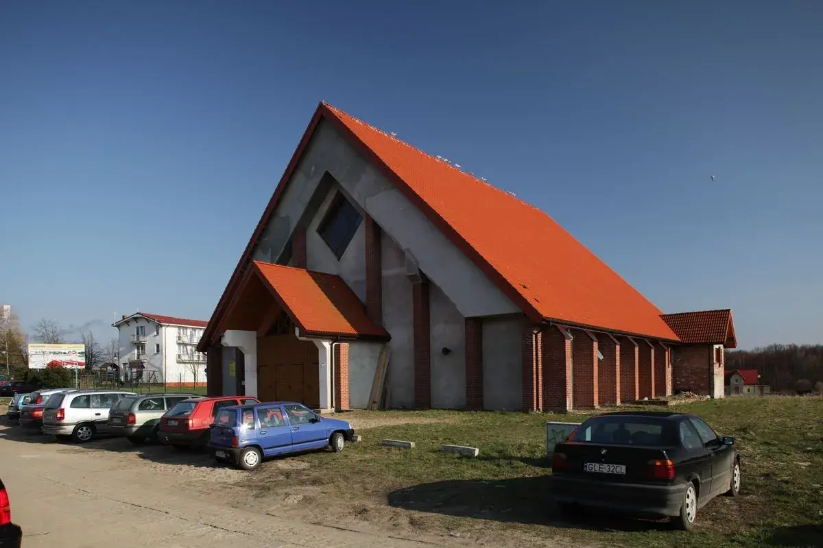 Nowoczesny kościół w Jarosławcu z pomarańczowym dachem, otoczony parkingiem z kilkoma samochodami.