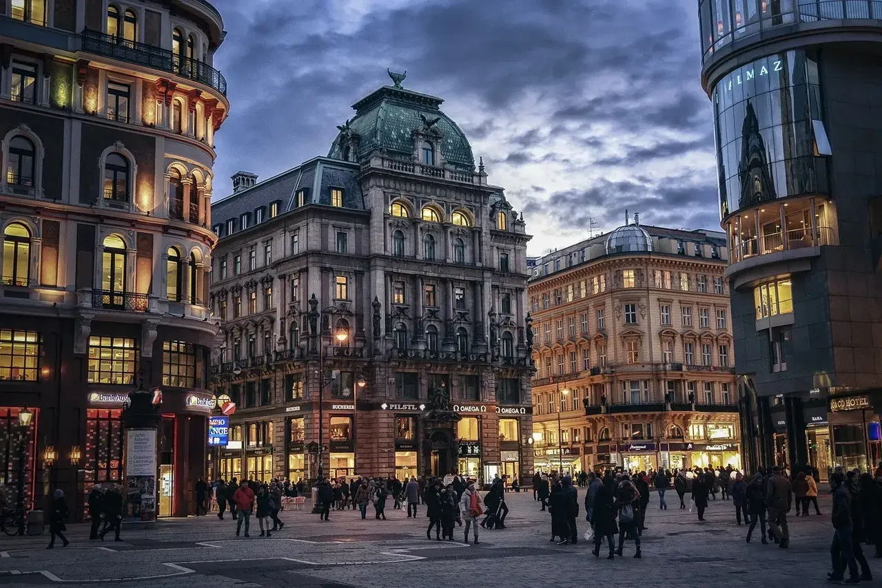 Wieczorny plac w Wiedniu, z pięknymi kamienicami i nowoczesnym budynkiem. Ludzie spacerują po oświetlonym deptaku, czuć atmosferę Wiednia Erdberg.