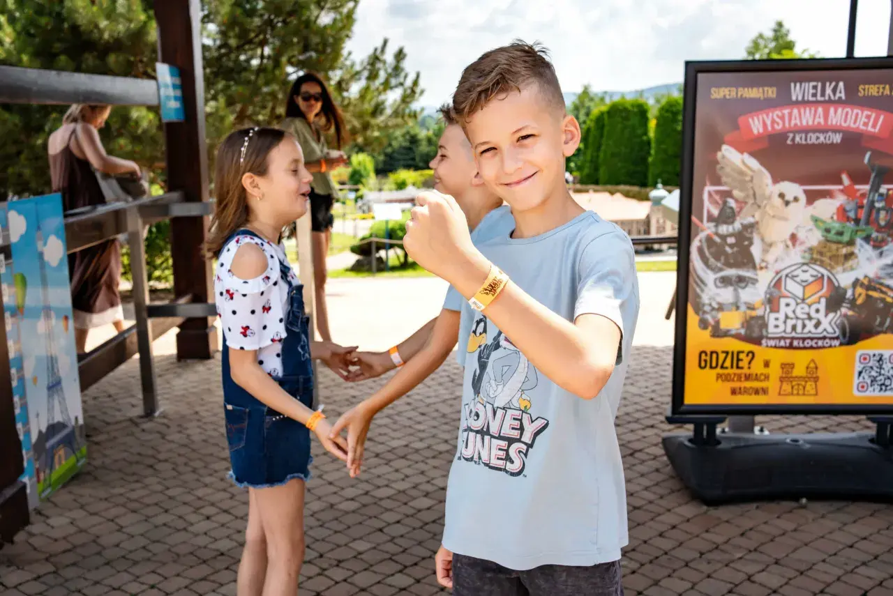 Uśmiechnięty chłopiec w parku rozrywki, obok niego dzieci. W tle plakat "Wielka wystawa modeli z klocków", który może zawierać informacje o **inwałd park cennik**.