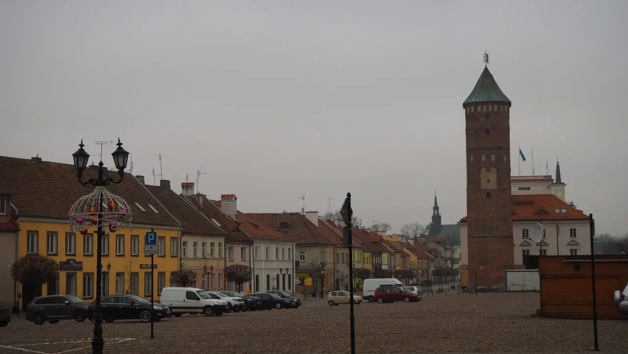 Rynek w Pułtusku z charakterystyczną wieżą. Kolorowe kamienice i zaparkowane samochody tworzą malowniczy widok.