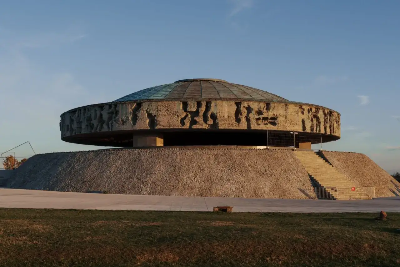 Monumentalne mauzoleum na terenie byłego obozu Majdanek. Kamienna podstawa i betonowy pierścień z płaskorzeźbami pod szklaną kopułą.