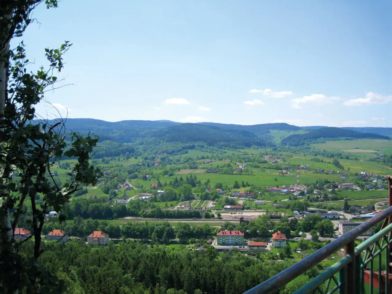 Widok na zieloną dolinę z zabudowaniami, w oddali wzgórza. Widać fragmenty szczytna zamek.