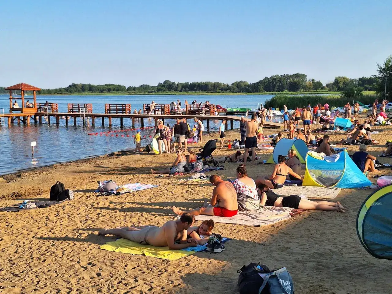 Plaża pełna ludzi, pomost, wodne atrakcje i słońce. Idealne miejsce na wypoczynek.