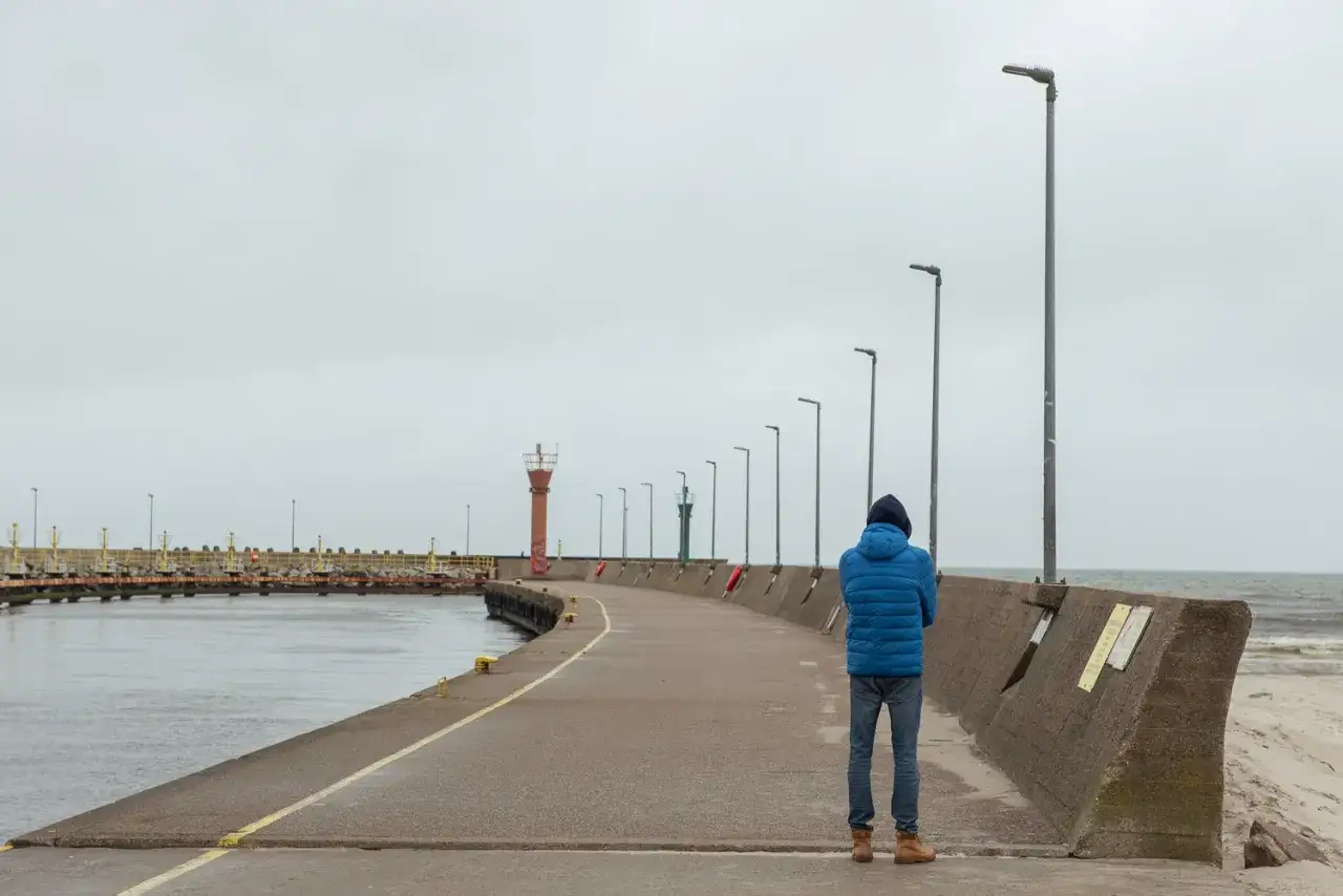 Mężczyzna w niebieskiej kurtce stoi na łebskiej promenadzie, patrząc na morze. W oddali widać latarnie morskie.