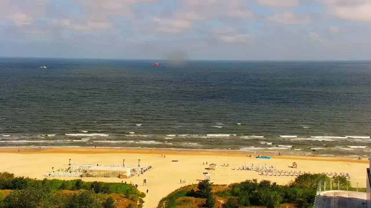 Słoneczny dzień na plaży w Świnoujściu. W oddali widać statek i czerwony spadochron. Deptak tętni życiem.