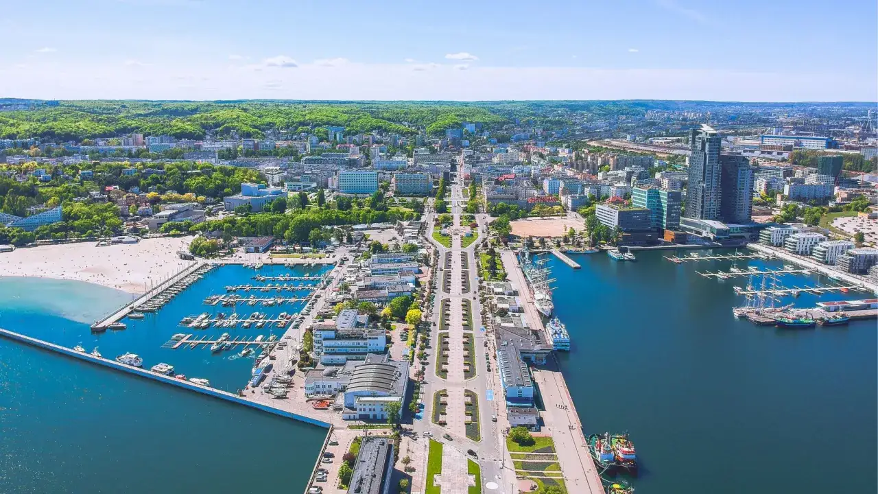 Gdynia Rynek Główny z lotu ptaka: szeroka aleja, marina pełna jachtów, plaża i nowoczesne budynki.