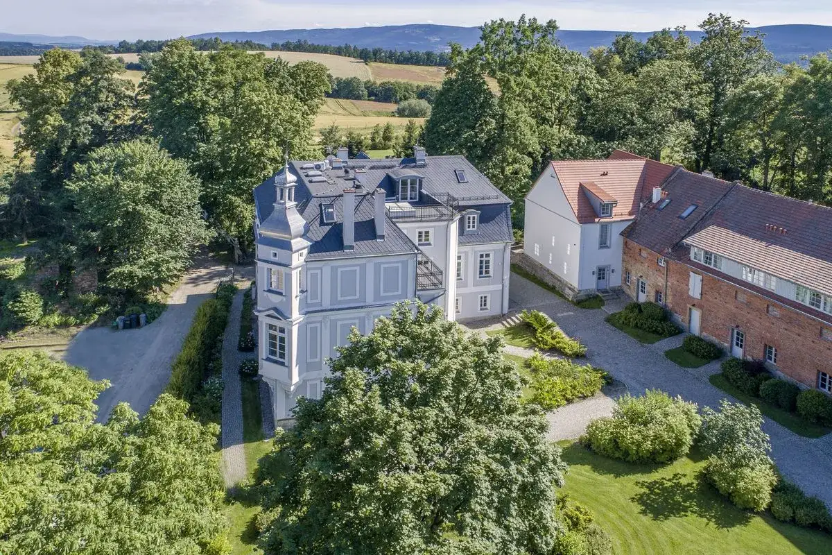 Elegancki dwór z wieżyczką, otoczony zielenią i polami, przypomina dawny kamieniec.