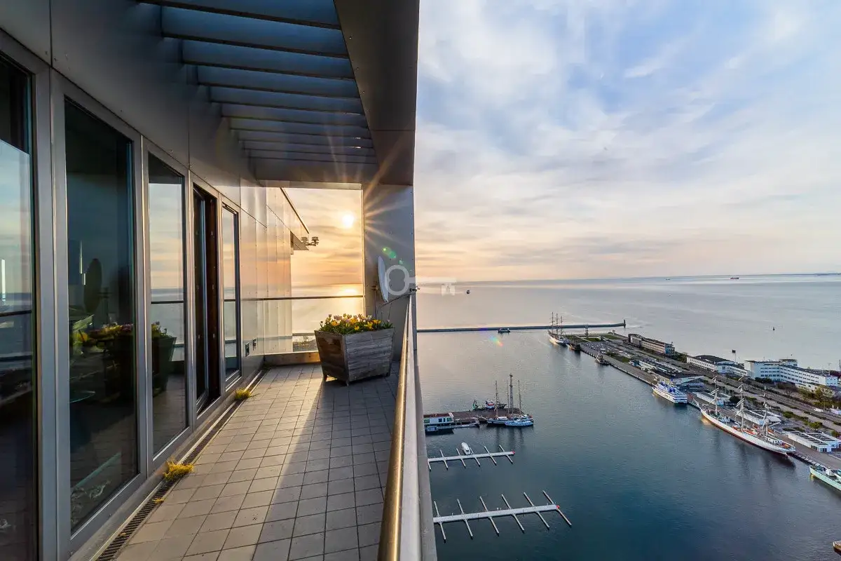 Widok z balkonu na port i morze o zachodzie słońca. W oddali widać żaglowce i inne statki, a także nowoczesne sea towers.