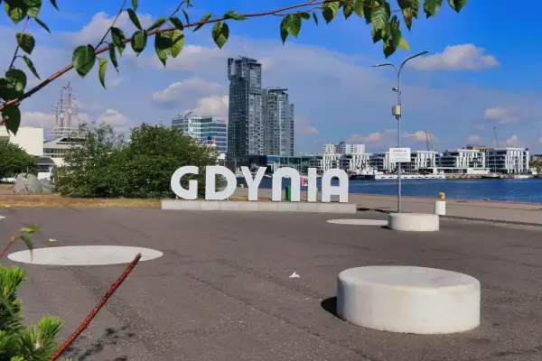 Napis "GDYNIA" na tle nowoczesnych budynków i portu. Widoczny żaglowiec i błękitne niebo.